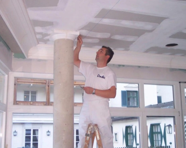 Ein Arbeiter streicht die Decke in einem hellen Raum mit großen Fenstern und einer Leiter.