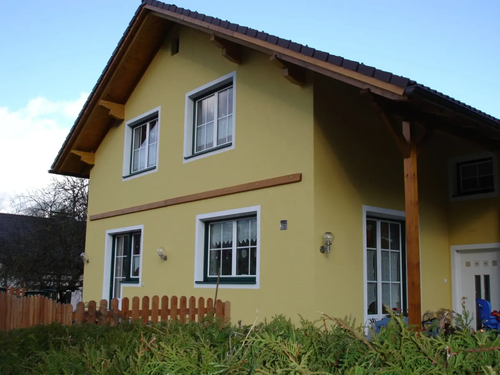 Gelbes zweistöckiges Haus mit weiß umrandeten Fenstern, braunem Dach und kleinem umzäunten Garten unter blauem Himmel