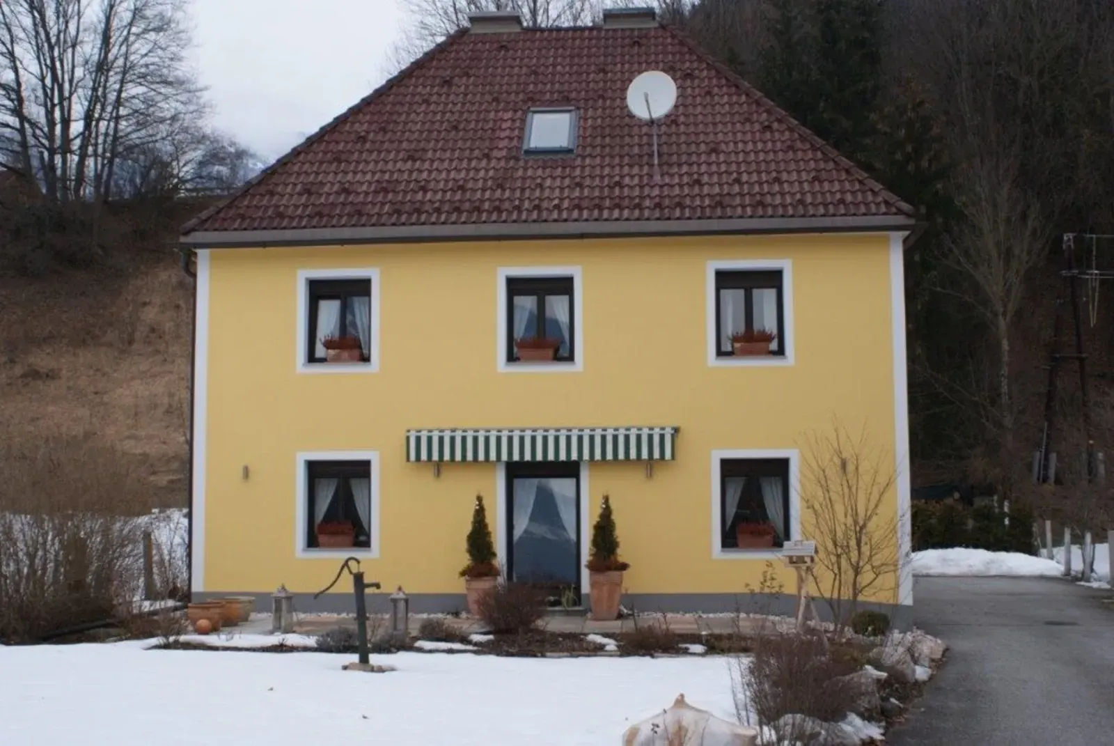 Gelbes Haus mit rotem Dach in verschneiter ländlicher Umgebung, Vordereingang mittig platziert, dahinter kahle Bäume.