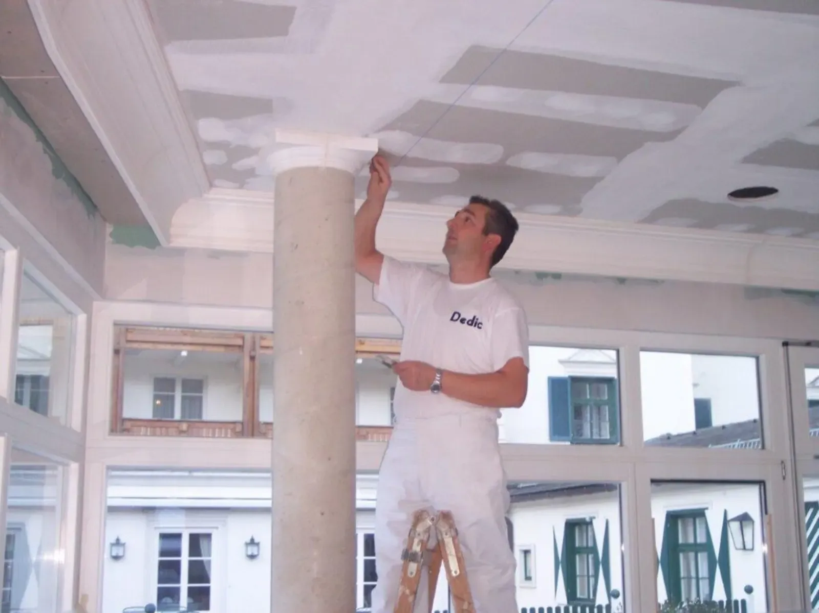 Ein Arbeiter verputzt die Decke in einem hellen Raum mit großen Fenstern.