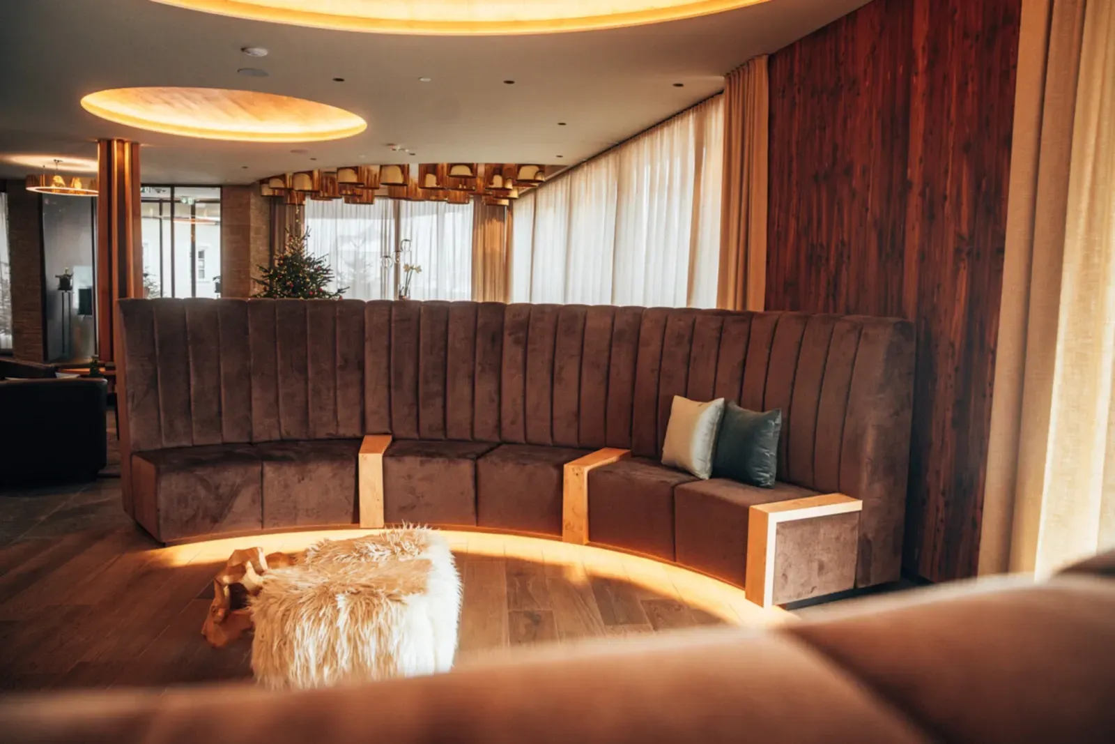Gemütliche, moderne Lounge mit geschwungenem braunem Sofa, Holzvertäfelung, runden Deckenleuchten und einem flauschigen Hocker.