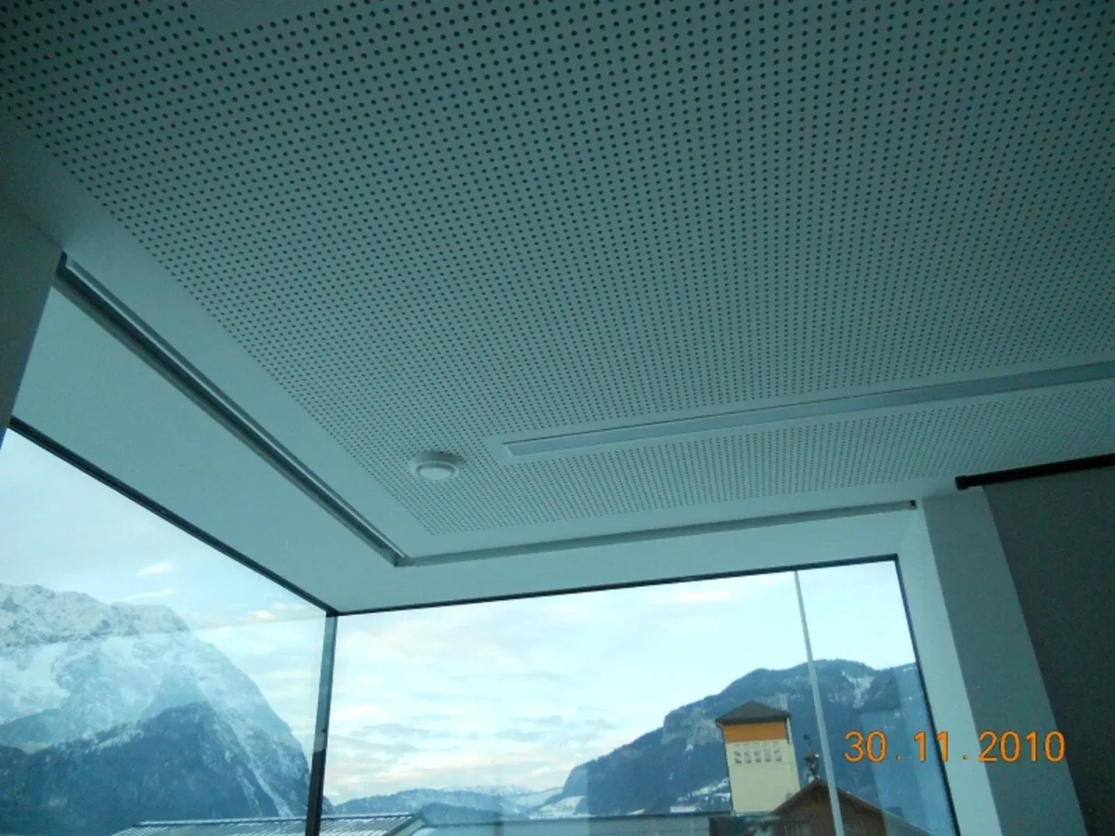 Blick auf die verschneiten Berge durch ein großes Fenster, mit gefliester Decke und einem kleinen Gebäude draußen.