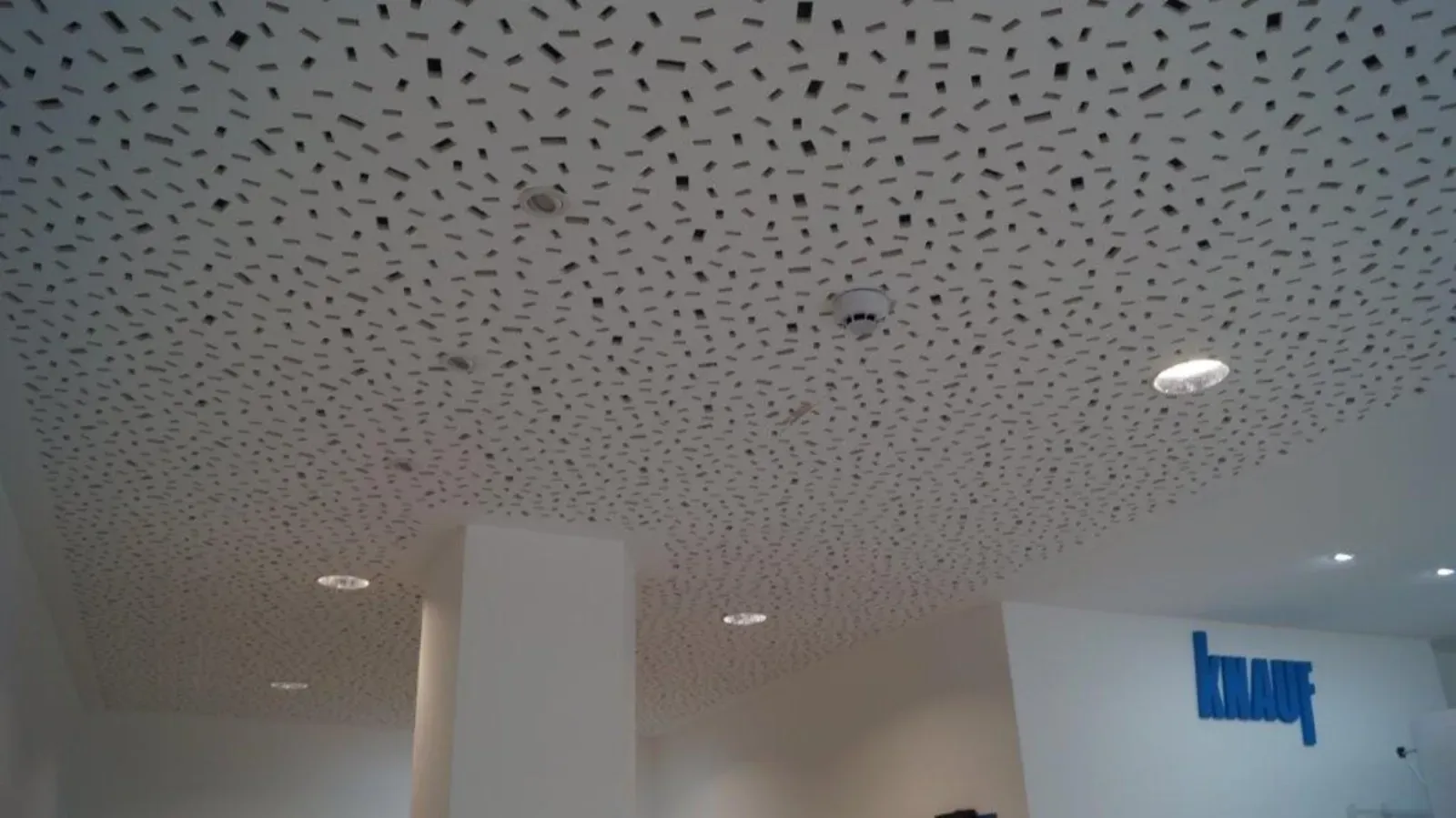 Weißer Innenraum mit gesprenkelter Decke, Einbauleuchten und einem blauen Wandlogo auf der rechten Seite.