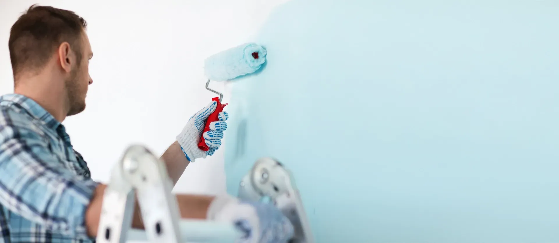 Ein Maler trägt mit einer Farbrolle hellblaue Farbe an eine Wand in der Nähe einer Leiter auf.
