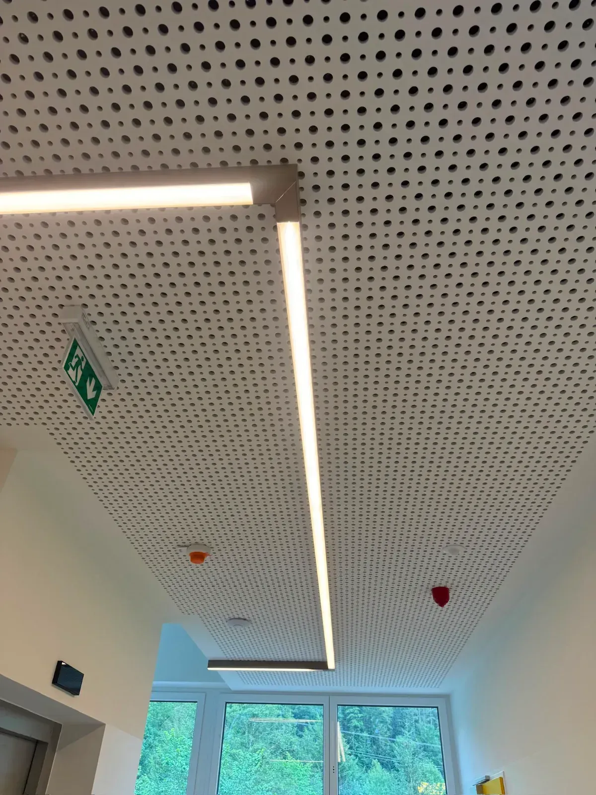 Decke mit perforierten Paneelen, linearen Leuchten und einem grünen Notausgangsschild über einem Flur.
