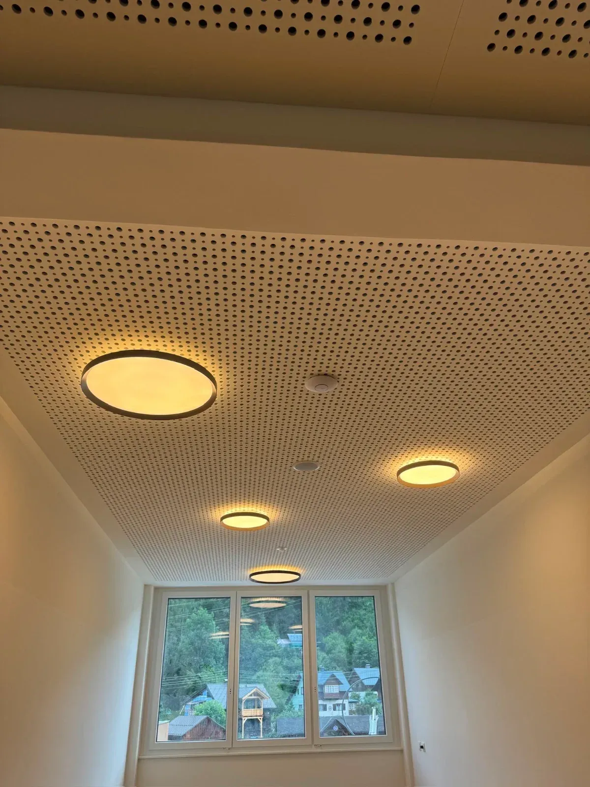 Weiße, perforierte Decke mit eingelassenen Leuchten über einem Zimmer mit Fenstern.