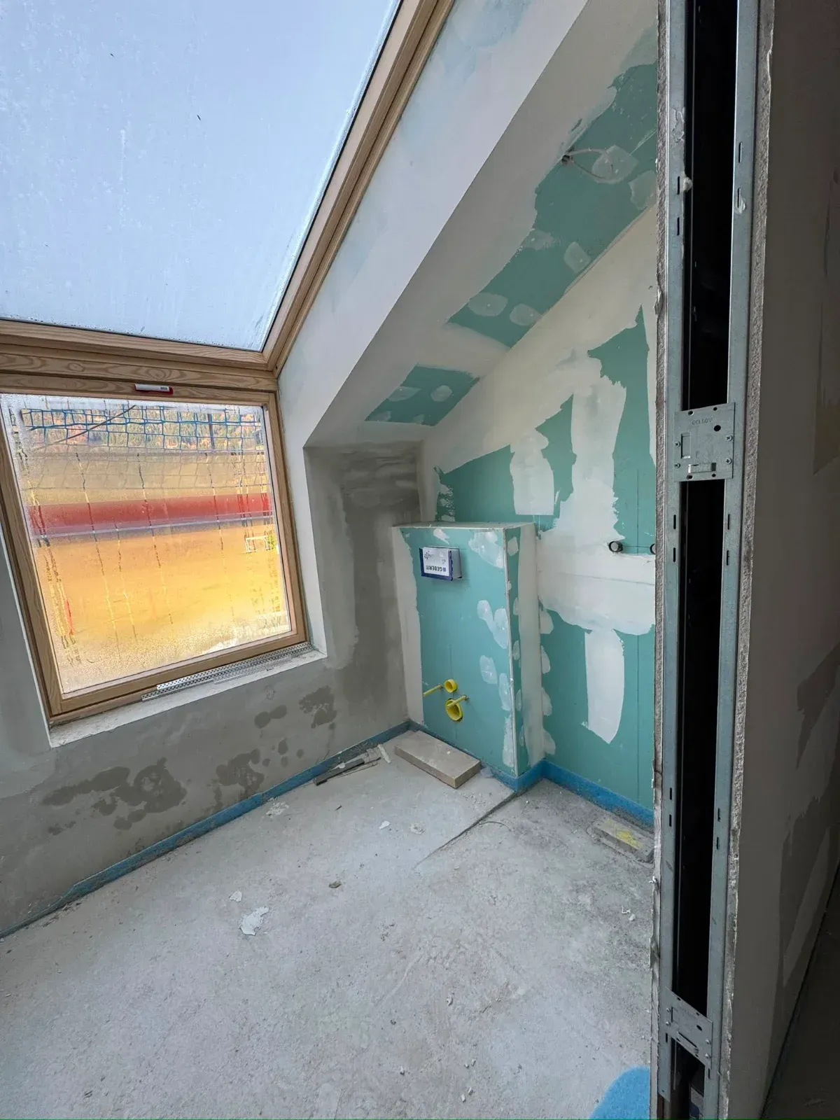 Kleiner, unfertiger Raum mit schrägem Fenster, blauen Schränken und Trockenbau-Ausbesserungen an den Wänden.