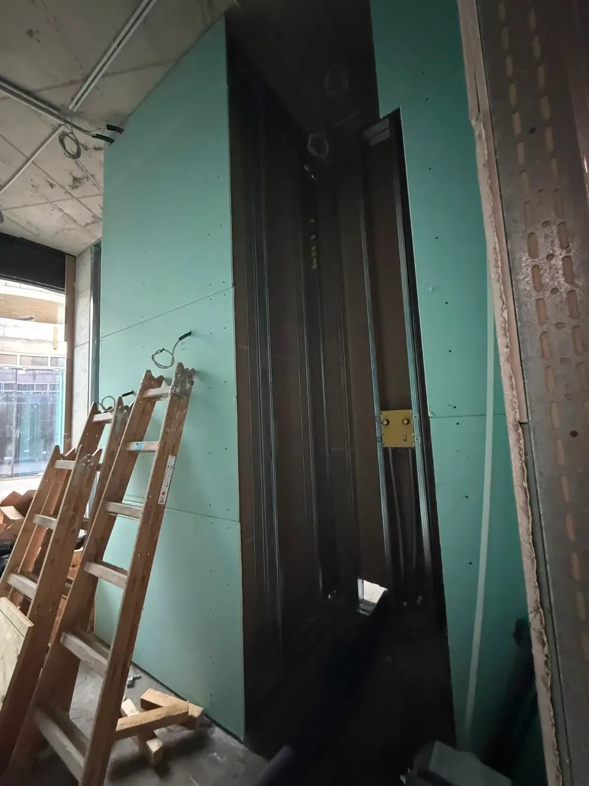Eine Holzleiter lehnt an einer türkisfarbenen Wand neben einem schmalen, dunklen Türrahmen in einem unfertigen Innenraum.