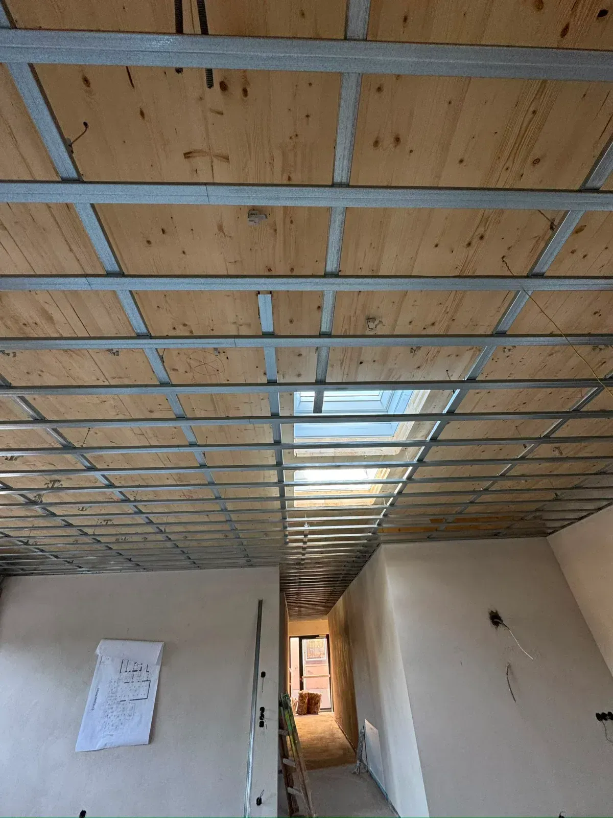 Ein leerer, im Bau befindlicher Raum mit freiliegendem Holzdeckengitter und einem hellen Türdurchgang am anderen Ende.