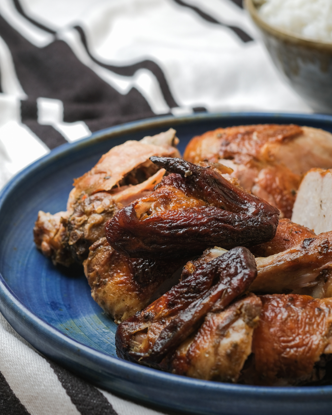 Pollo asado en un plato azul, con un tazón de arroz al fondo. Tela a rayas blancas y negras.