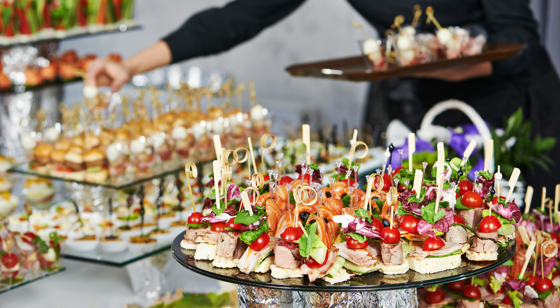 Buffet con entremeses en bandejas de varios niveles; una persona sirve más aperitivos.