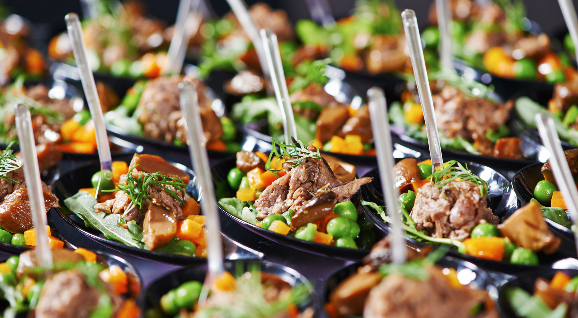 Pequeños aperitivos en bandejas negras, cubiertos con carne, verduras y hojas verdes, sujetos con brochetas transparentes.