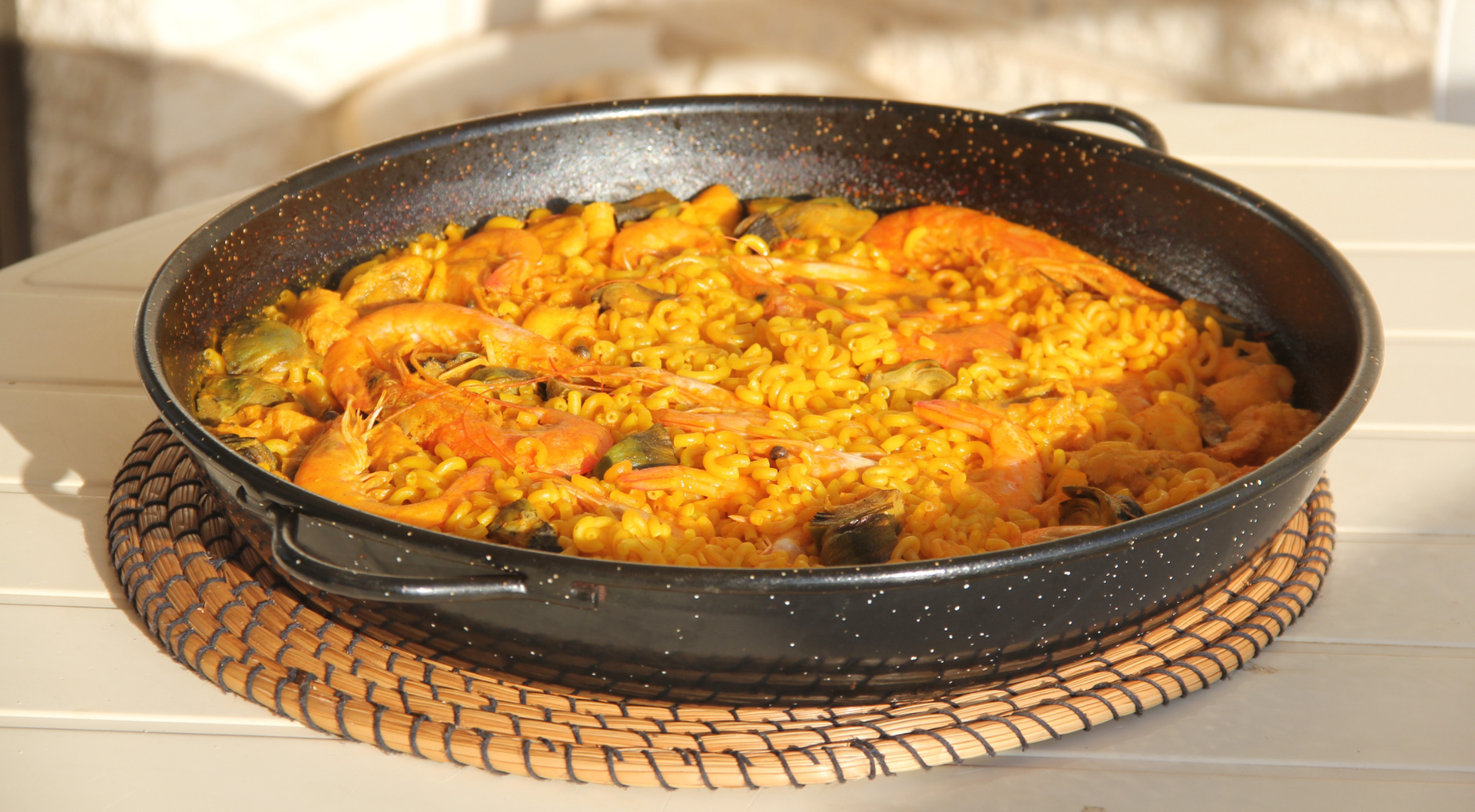 Paella en paellera negra, con gambas, sobre una estera tejida en un entorno al aire libre.