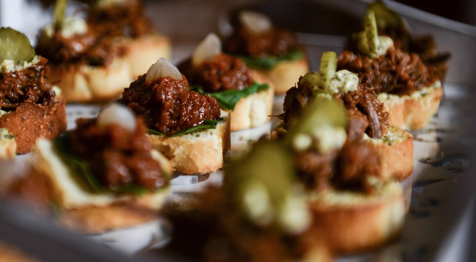 Aperitivos sobre pan tostado cubiertos con carne, pesto y pepinillos en una bandeja.