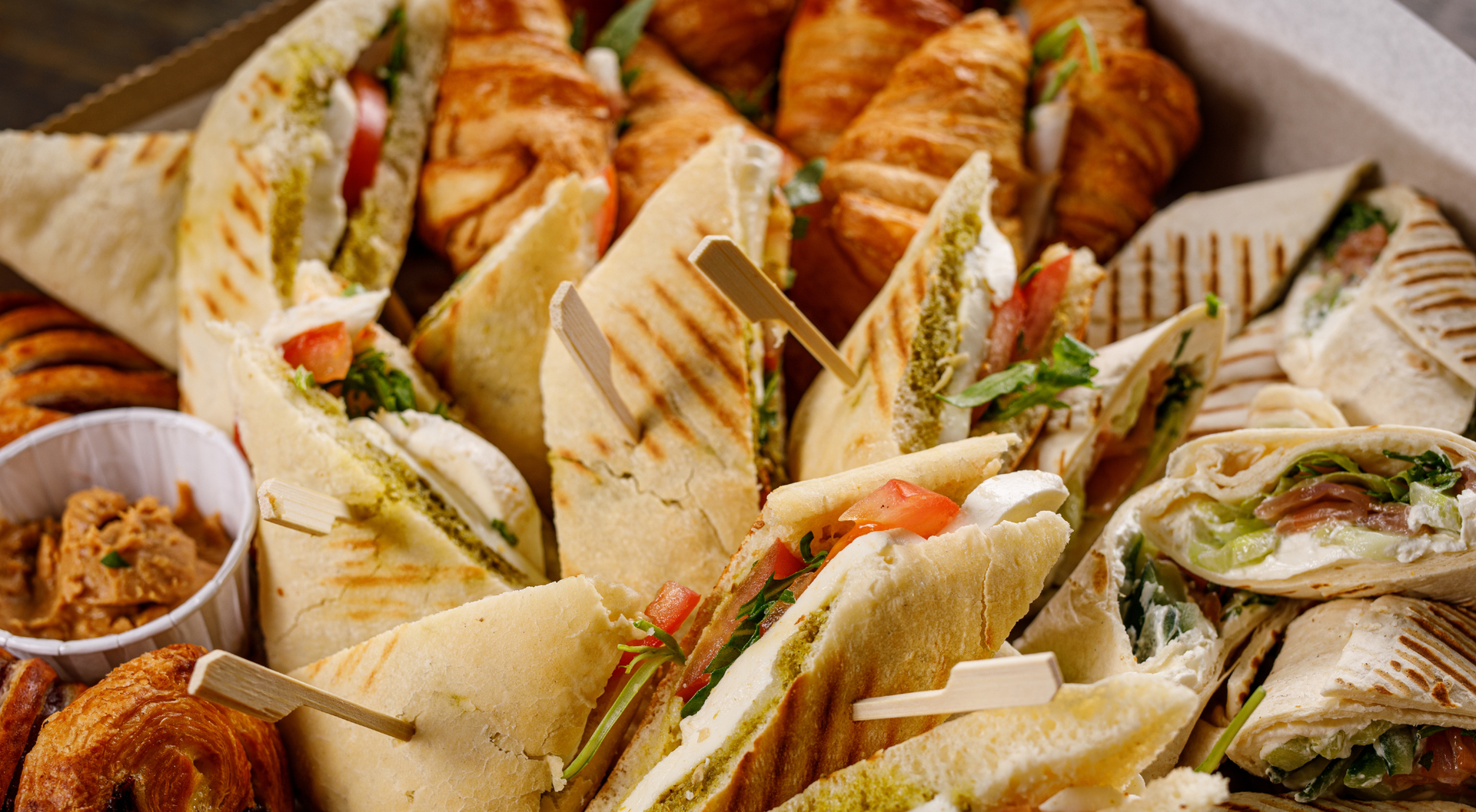 Surtido de sándwiches y pasteles en caja, incluyendo wraps y croissants, con una salsa para untar.