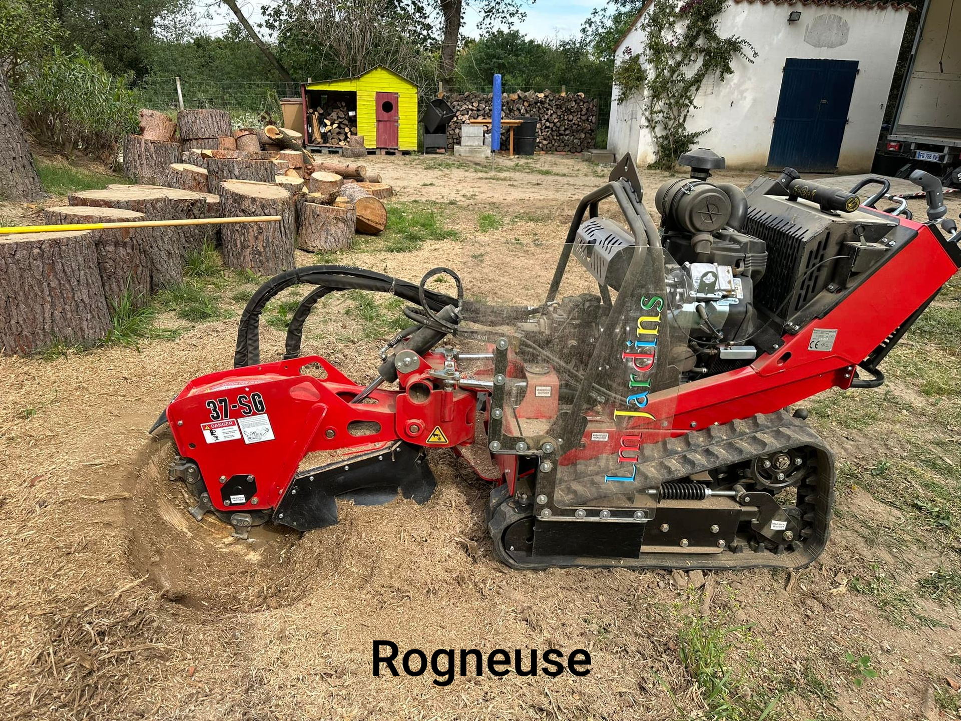 Broyeur de souches rouge sur chenilles, en train de broyer une souche dans une cour. Un tas de bois et un petit abri se trouvent à l'arrière-plan.