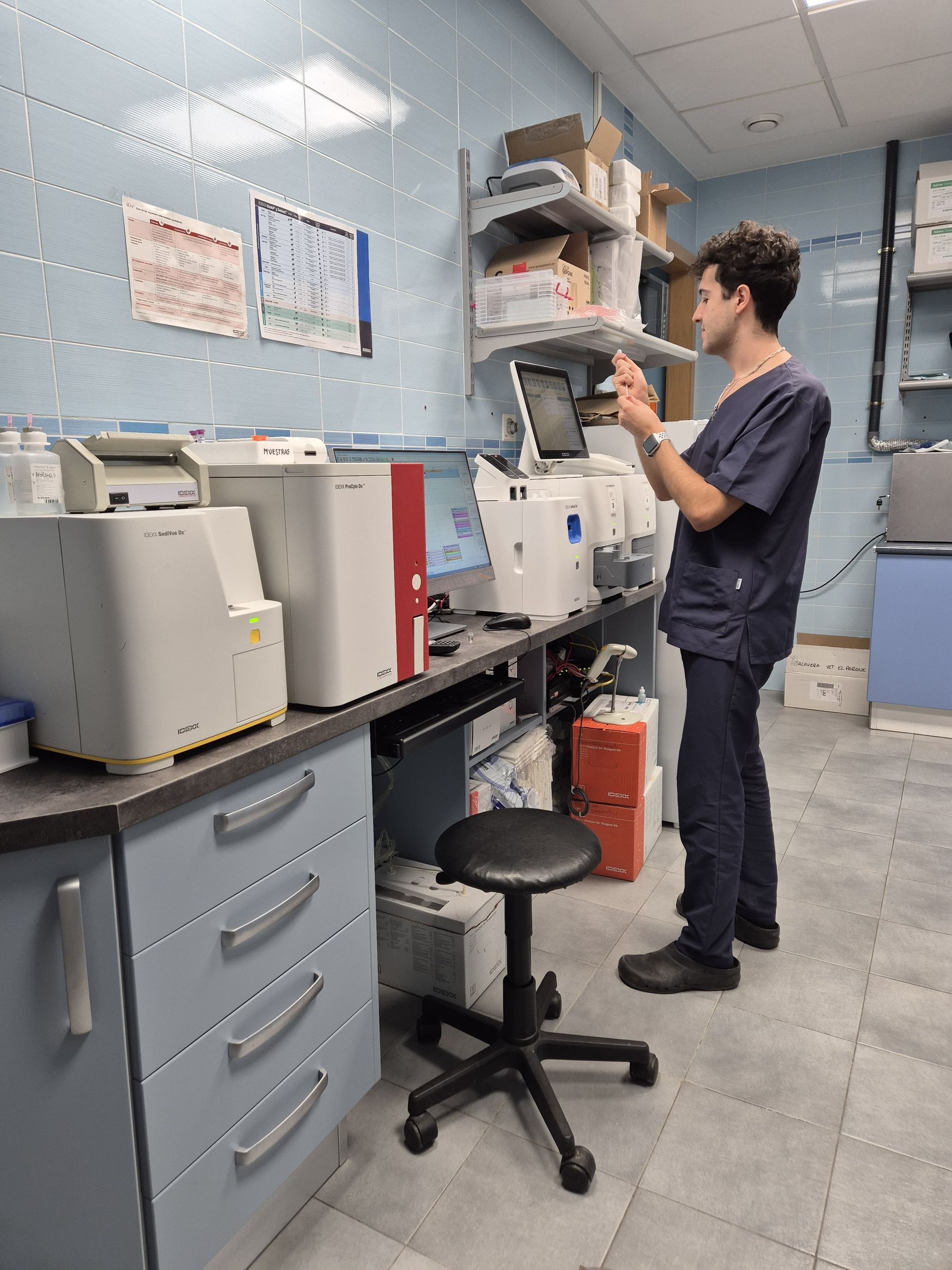 Un hombre está de pie en un laboratorio con varias máquinas para la realización de analíticas sanguíneas y otros fluidos, un ordenador en la encimera y un frigorífico en una esquina.