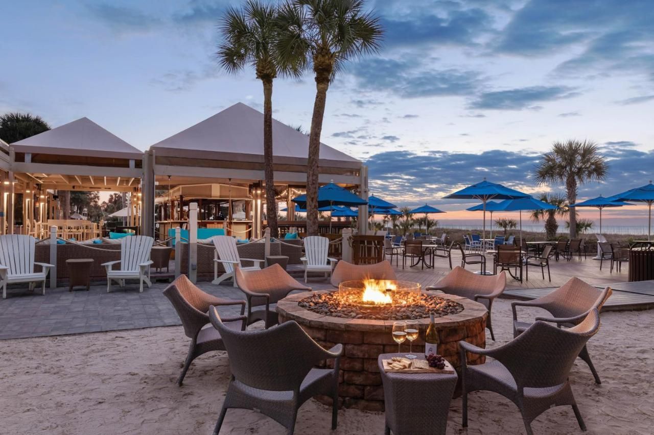 A fire pit is surrounded by chairs and tables on the beach.