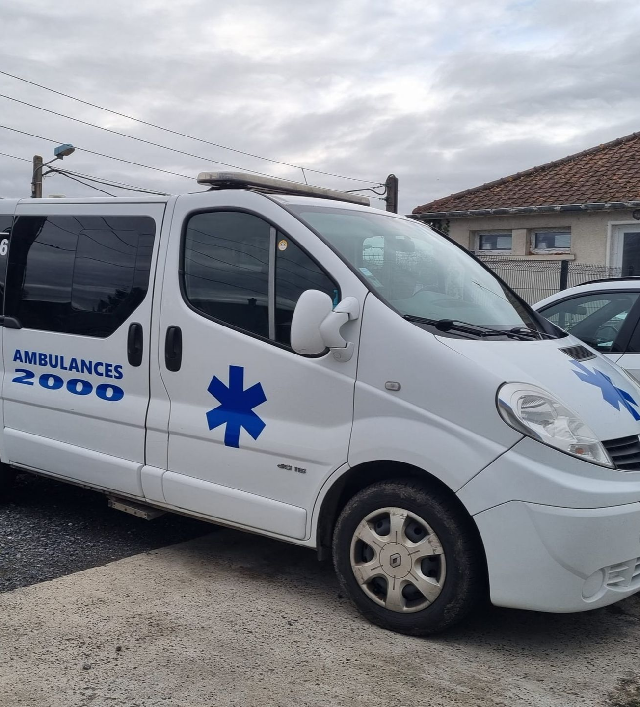 Une ambulance blanche ornée d'une étoile de vie bleue et portant l'inscription Ambulances 2000 est stationnée devant un bâtiment.