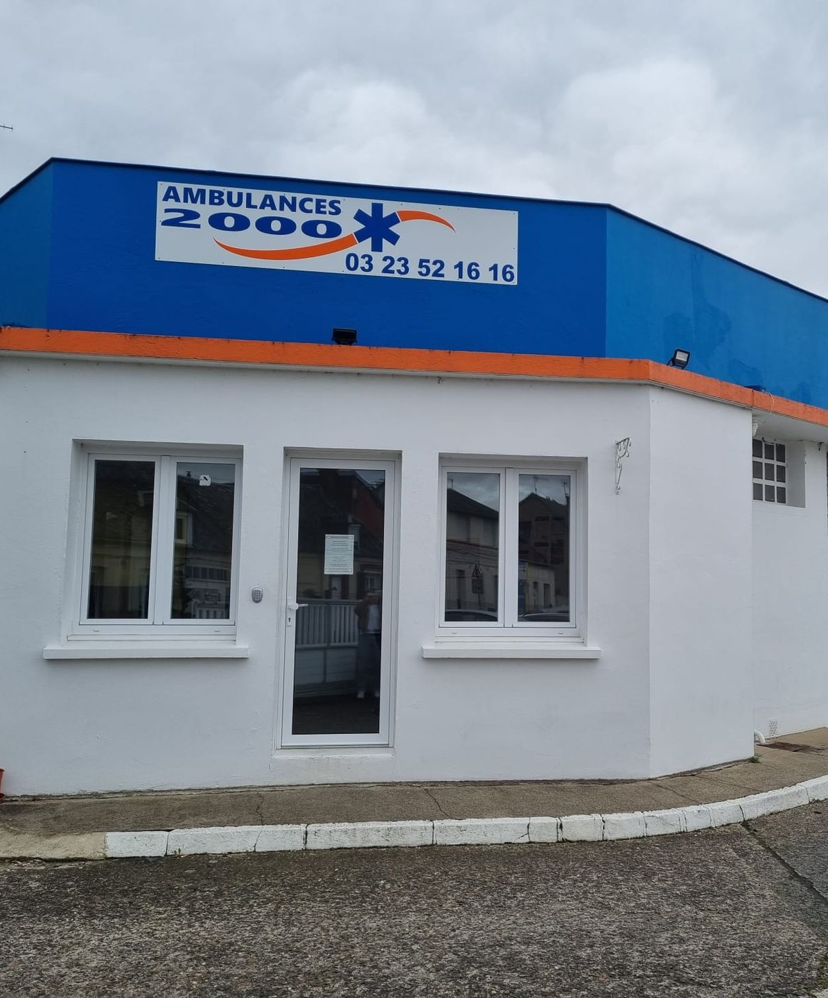 Bâtiment blanc avec un panneau bleu et orange de l'enseigne Ambulances 2000 et un numéro de téléphone.