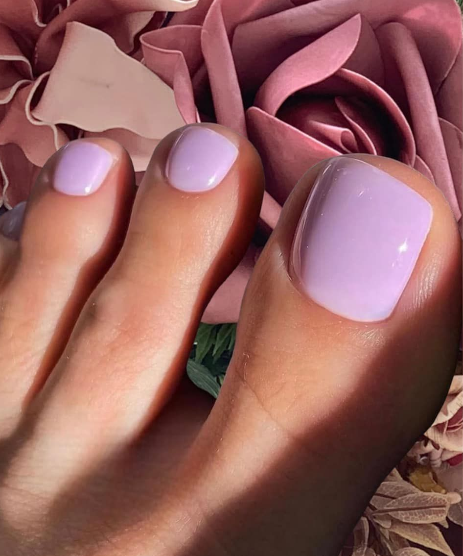 Primer plano de un pie con esmalte de uñas de color lila, rodeado de pétalos de rosa rosa.
