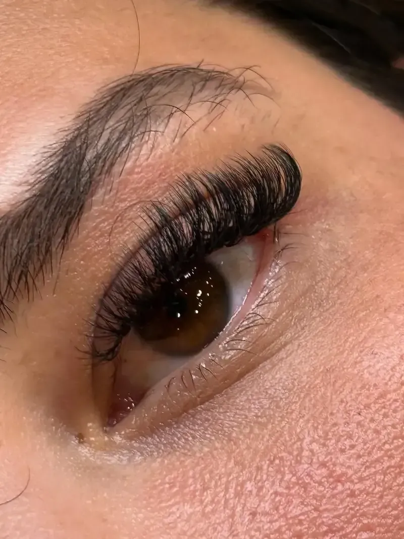 Primer plano de un ojo con extensiones de pestañas oscuras y gruesas. Un ojo marrón está abierto y la ceja está arqueada.