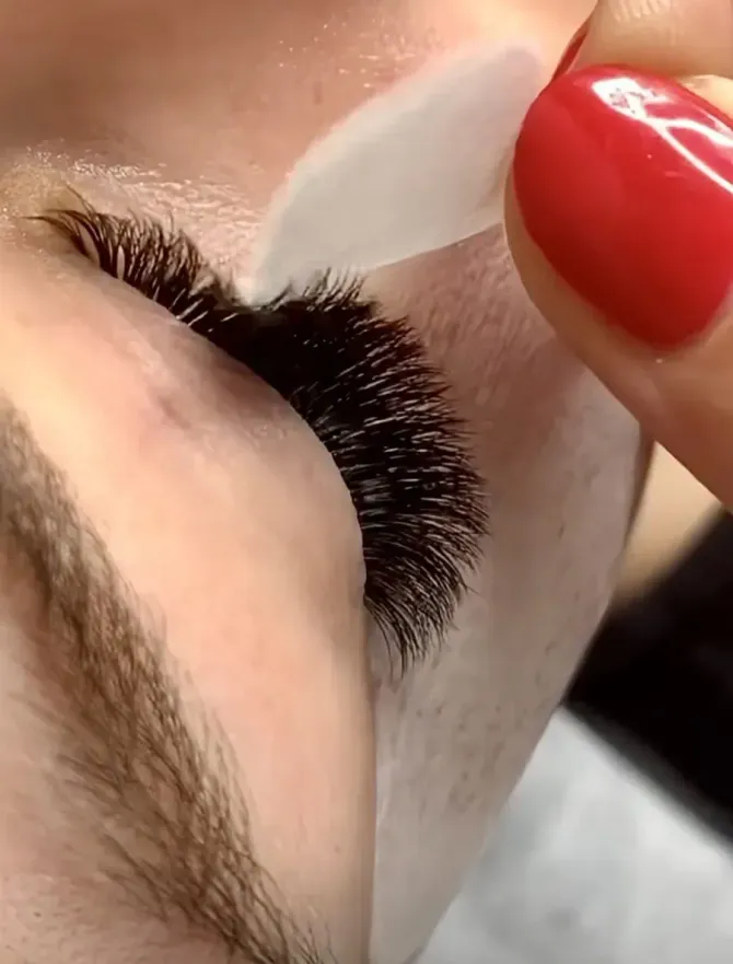 Primer plano del ojo de una persona con extensiones de pestañas gruesas y oscuras; los dedos con uñas rojas