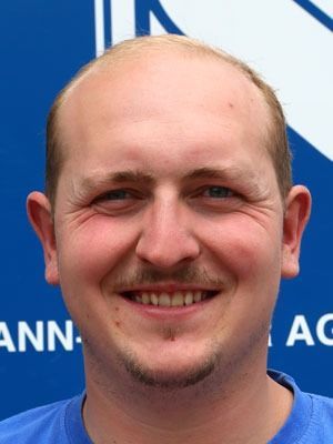 Lächelnder, glatzköpfiger Mann mit Schnurrbart und Spitzbart in einem blauen Hemd vor blauem Hintergrund
