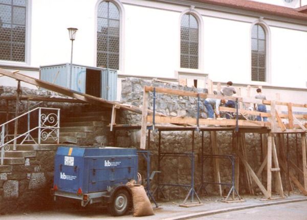 Ein blauer Baucontainer und ein Baugerüst stehen neben einem Gebäude mit Bogenfenstern und einer Steinmauer.