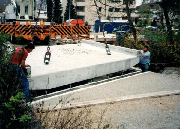 Arbeiter bewegen eine grosse weisse Betonplatte mit einem Kran auf einer abschüssigen Strasse in der Nähe von orangefarbenen Absperrungen.