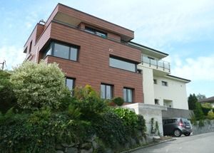 Modernes mehrstöckiges Haus mit roten Ziegeln und weisser Verkleidung an einer abfallenden Strasse, teilweise von Grün verdeckt