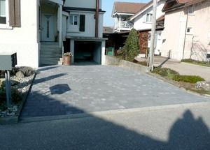 Wohnhausauffahrt mit grauen Pflastersteinen zwischen weissen Häusern, von der Strasse aus gesehen.