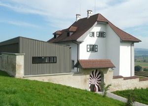 Weisses Haus mit braunem Dach neben einem grauen, scheunenartigen Gebäude in einer grasbewachsenen ländlichen Landschaft
