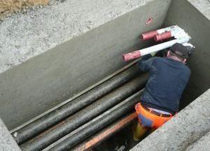 Ein Arbeiter hantiert in einem Betongraben mit Rohren; rote Rohrenden sind sichtbar, und er trägt eine orangefarbene Sicherheitshose.