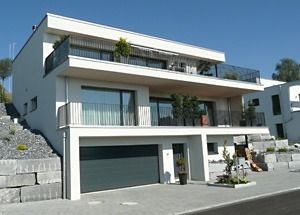 Modernes, weisses, mehrstöckiges Haus mit Balkonen, Garage und Steingartenanlage an einem sonnigen Hang