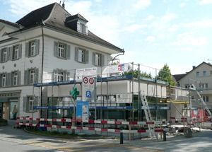 Weisses Haus mit Baugerüsten und Baustellenabsperrungen an einer Strassenecke unter einem teilweise bewölkten Himmel