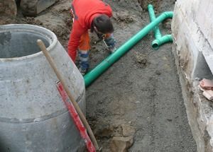 Ein Arbeiter in Rot beugt sich über einen Graben neben grünen Rohren und einem Betonring.