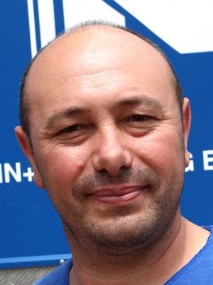 Lächelnder, glatzköpfiger Mann in einem blauen Hemd vor einem blauen Schild
