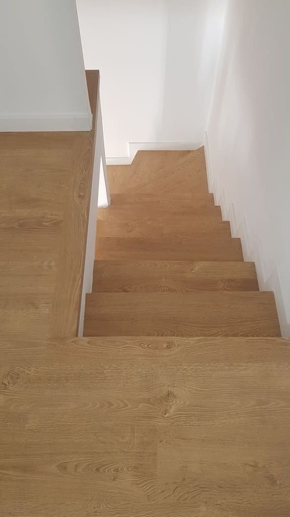 Escalera de madera que desciende junto a una pared blanca, vista desde el rellano superior.