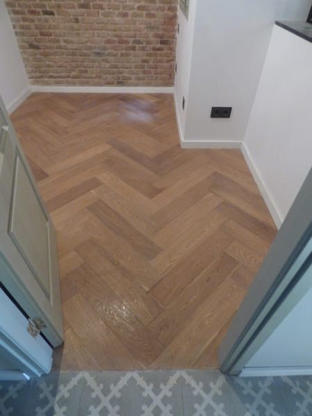 Se abre una puerta que da a una pequeña habitación con suelo de madera en espiga, pared de ladrillo y paredes blancas.