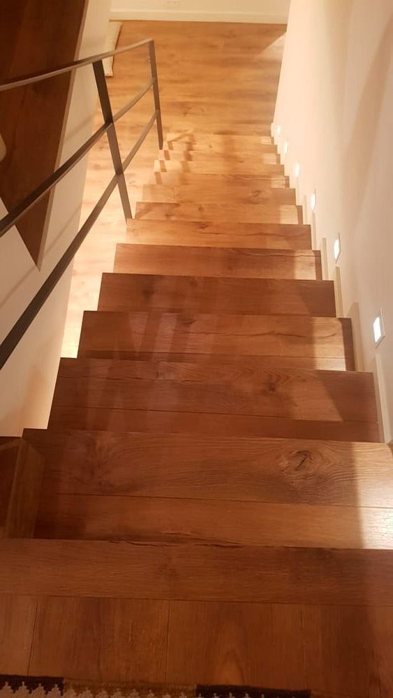 Escalera de madera que desciende hasta un rellano, con barandilla de metal negro e iluminación interior cálida.