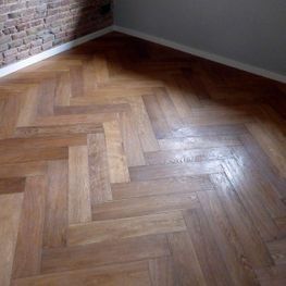 Rincón de habitación vacío con suelo de madera en espiga, paredes grises y molduras de ladrillo visto.