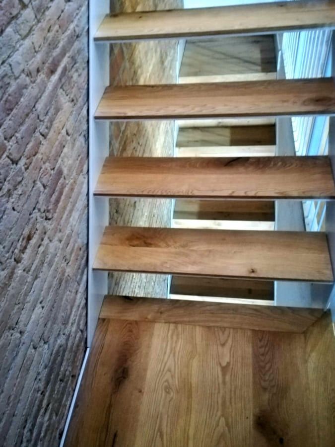 Escalera de madera con peldaños abiertos y una pared de ladrillo al lado.