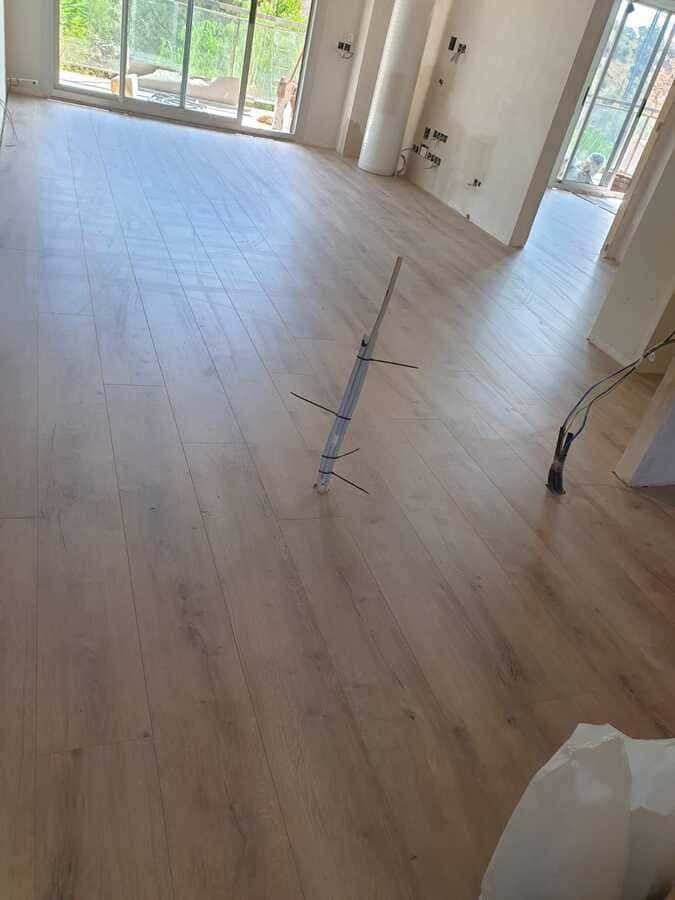 Habitación vacía con suelo de madera clara, grandes ventanales y cableado a la vista en el centro del suelo.
