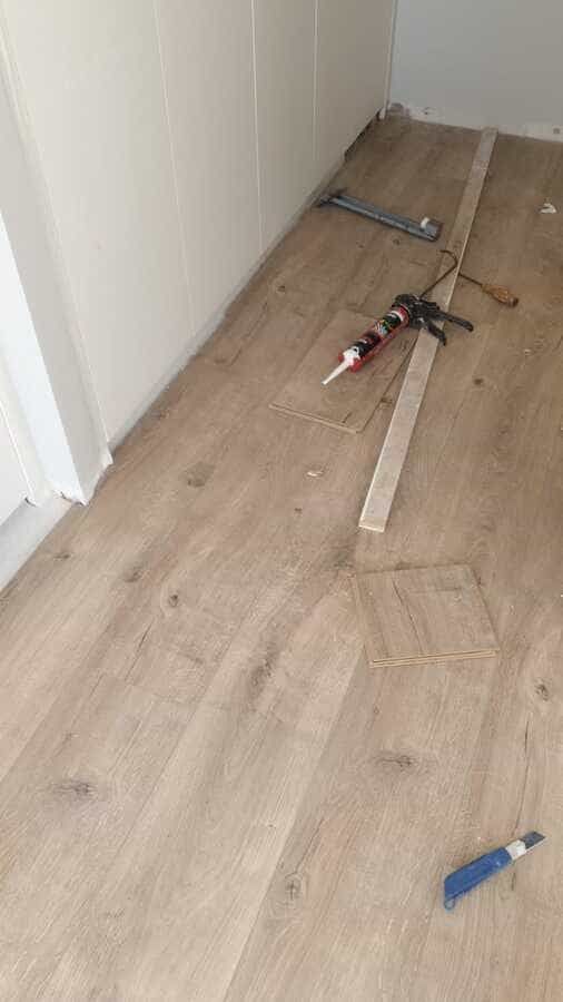 Suelo de madera junto a una pared blanca con herramientas, listones de madera y escombros esparcidos por el suelo.