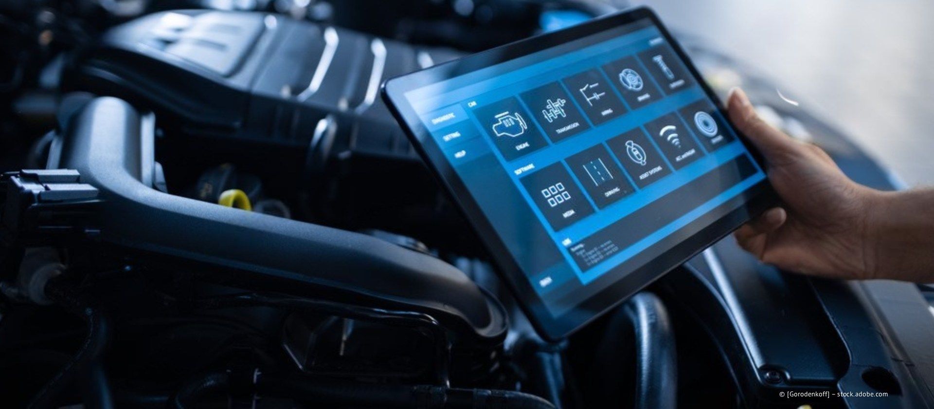Eine Hand hält ein Tablet mit Fahrzeugdiagnosedaten neben dem Motorraum.