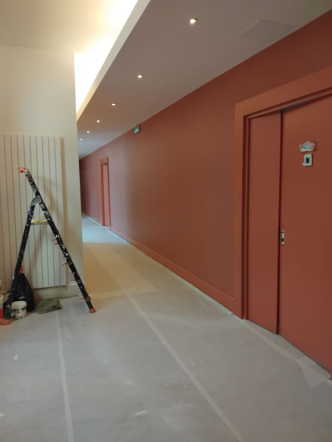 Long couloir avec des murs et des portes de couleur corail, un plafond blanc et un escabeau.