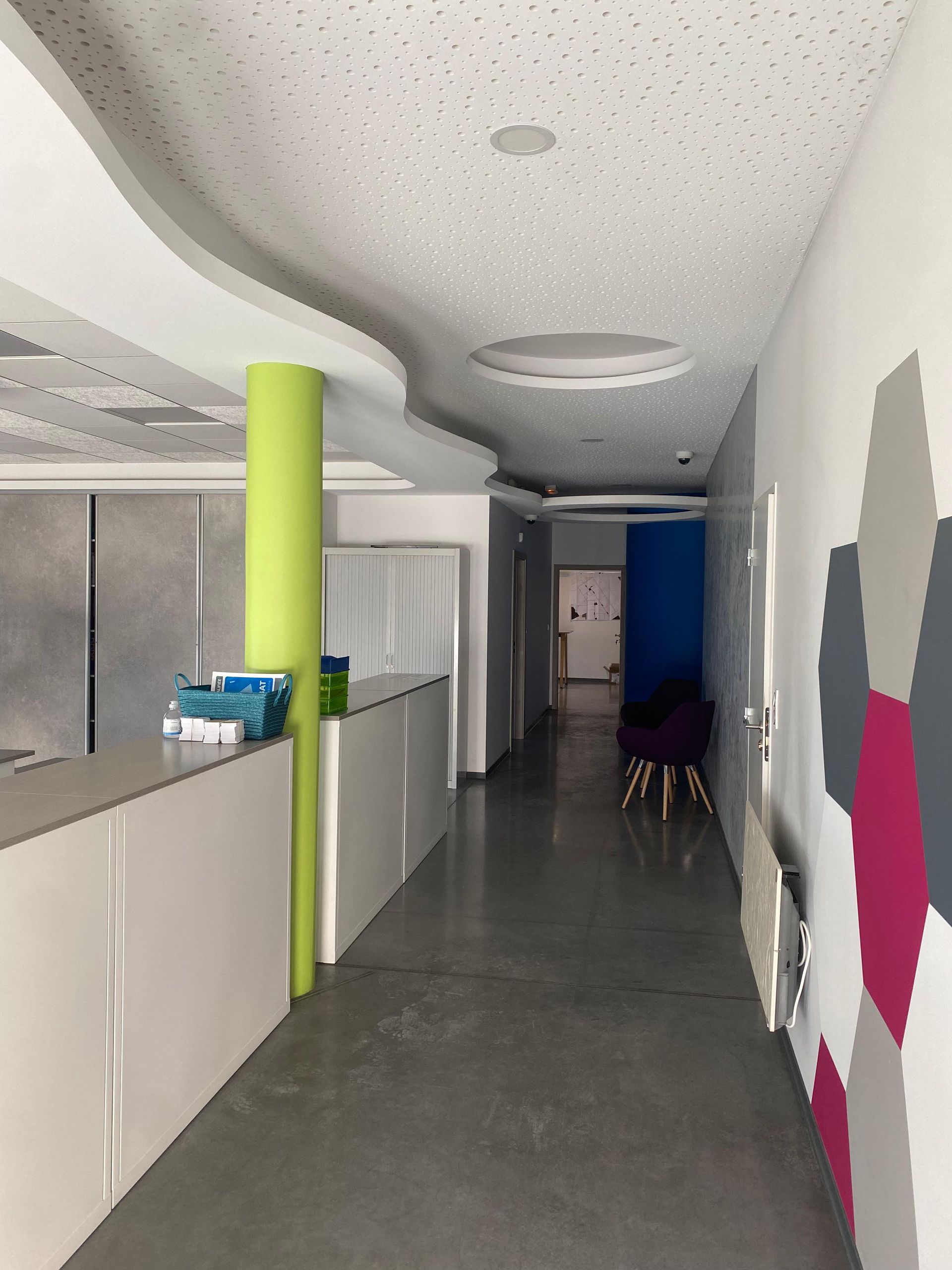 Long couloir avec des murs blancs et gris, un pilier vert et un bureau de réception.
