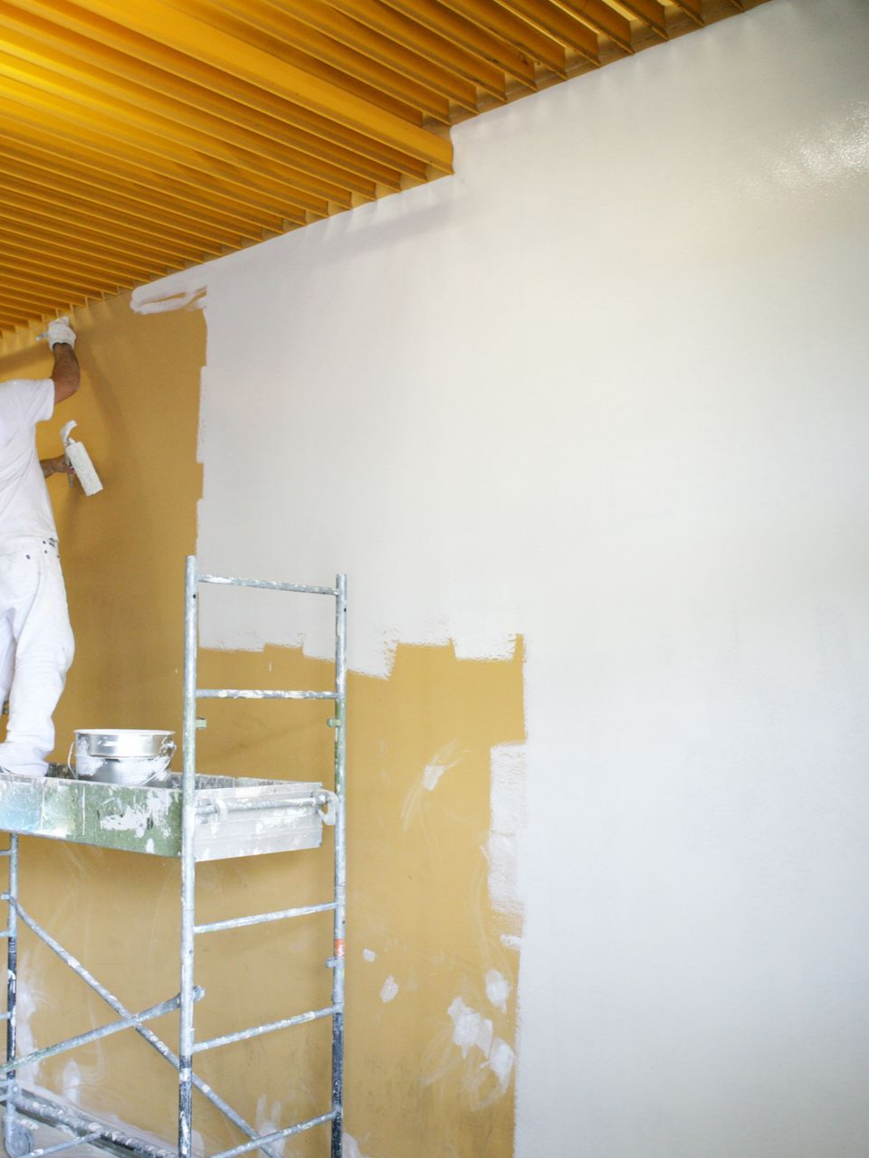 Une personne sur un échafaudage peint un mur en blanc, à côté d'un plafond jaune.