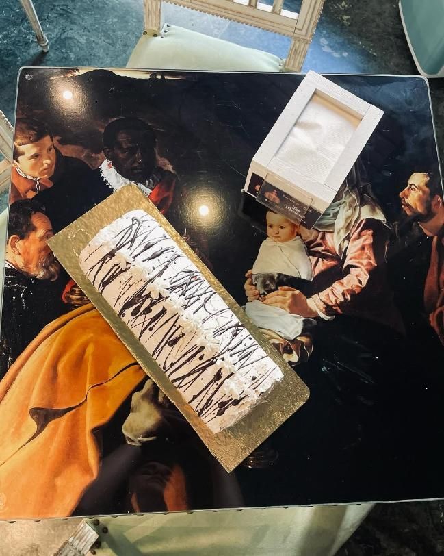 Pastel en bandeja de oro y caja de pañuelos sobre una mesa con pintura de figuras.